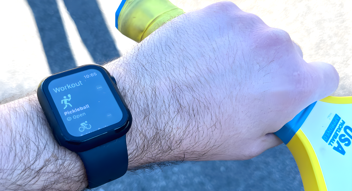 Detail of an Apple watch displaying a pickleball workout feature while the hand holds a pickleball paddle