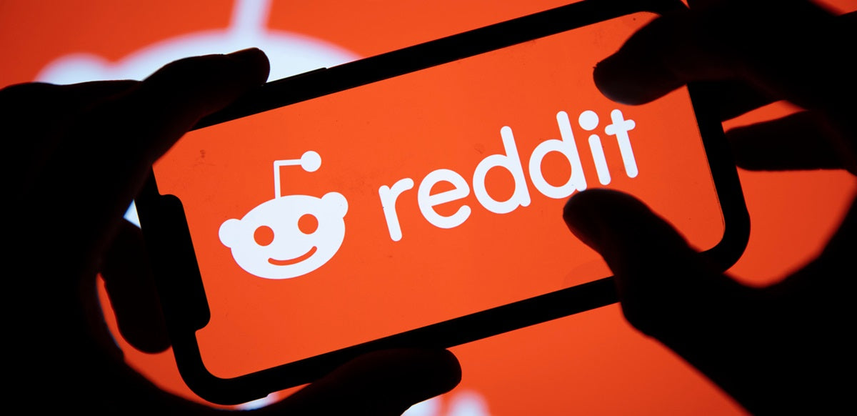 A view of Reddit's logo as viewed full-screen on a phone, with hands holding the phone