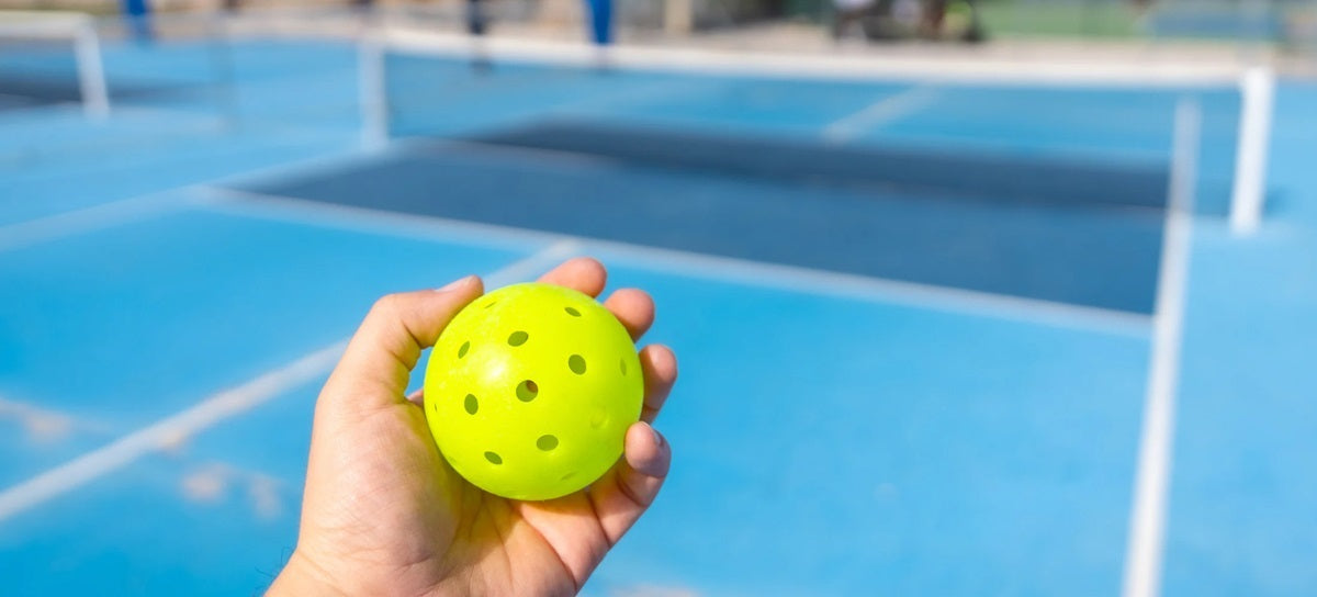 An isolated hand holding a pickleball with a court in the background